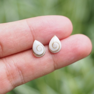 Peut inclure: Une paire de boucles d'oreilles en forme de goutte d'eau. Chaque boucle d'oreille est blanche avec un motif en spirale marron. Les boucles d'oreilles sont tenues dans la main d'une personne sur un fond vert flou.