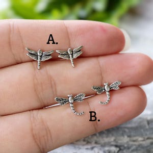 May include: Four silver-toned dragonfly-shaped stud earrings arranged in two pairs labelled A and B. Pair A features dragonflies with wings spread horizontally, while pair B features dragonflies with curved tails. Each piece has detailed wing textures.