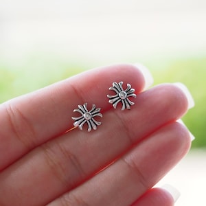 Op de afbeelding: Een paar kleine, zilverkleurige oorstekers. Elke oorbel heeft een gestileerd, symmetrisch bloemmotief met een centraal, cirkelvormig accent. De oorbellen worden getoond tegen een zachte, vage achtergrond.