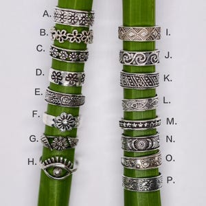 Può includere: Una collezione di sedici anelli in metallo color argento etichettati da A a P, con vari motivi in rilievo tra cui fiori, forme geometriche, onde, stelle, motivi celesti e un design a occhio, disposti su due steli verdi verticali.