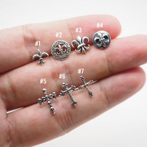 May include: A collection of silver-toned stud earrings in various designs. The earrings include fleur-de-lis, circular patterns, and cross shapes. The image shows seven different styles, each with a unique design, suitable for multiple piercings or styling options.