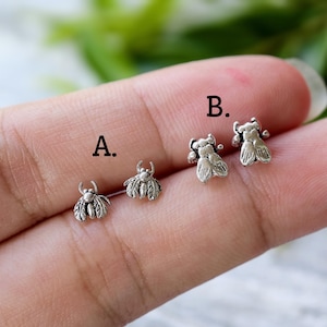 May include: Two pairs of small silver-toned insect-shaped stud earrings. The pair labelled A features a detailed beetle design, while the pair labelled B shows a fly-like insect. Both sets have a textured, oxidised finish with visible wings.