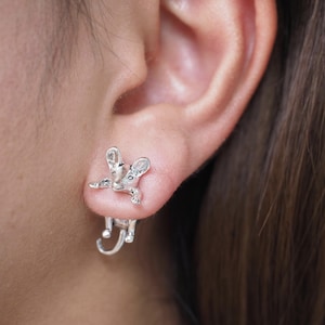 Peut inclure: Boucles d'oreilles argentées en forme de souris. Les boucles d'oreilles sont conçues pour donner l'impression qu'une souris grimpe sur l'oreille. La souris présente des détails tels que les oreilles, les yeux et les pattes. Un accessoire unique et fantaisiste.