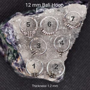 Pode incluir: Uma coleção de brincos de argola Bali em prata, cada um com designs decorativos únicos. Os brincos têm 12 mm e 1,2 mm de espessura. O texto "12 mm Bali Hoop Earrings" e "Thickness: 1.2 mm" são exibidos.