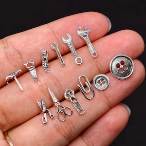 May include: A collection of small, silver-colored charms in various shapes, including tools like a hammer, saw, and wrenches, along with a paperclip, scissors, buttons, and a zipper. The charms are displayed against a neutral background.