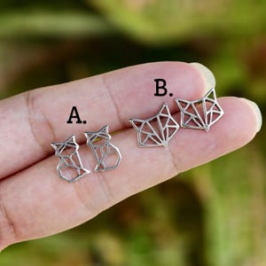 May include: Two pairs of silver-toned metal stud earrings featuring geometric fox designs. Pair A shows a sitting fox silhouette, while pair B displays a fox head outline. Both sets are labeled with black letters A and B for identification.