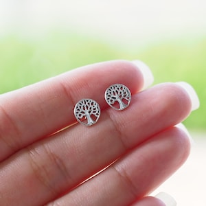 May include: A pair of small, silver-colored stud earrings. Each earring features a detailed tree design within a circular frame. The earrings are shown against a blurred green and white background.