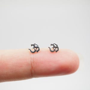 以下が含まれることがあります： 小さな銀色のOmシンボルスタッドピアス2つ。ピアスは明るい背景に配置されています。Omシンボルは、ヒンドゥー教、仏教、ジャイナ教における神聖な音と精神的なアイコンです。