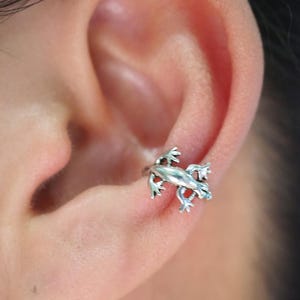 May include: A silver-colored frog ear cuff is shown on an ear. The frog is detailed with visible legs, feet, and a textured body. The cuff wraps around the ear's cartilage.
