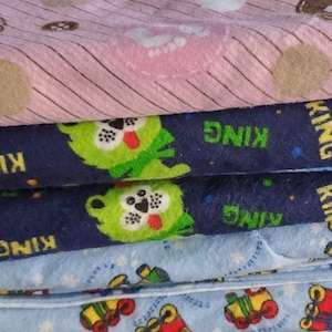 May include: A stack of five fabric squares with different patterns. The top square is pink with brown polka dots and stripes. The next square is blue with green cartoon bears and the word "KING" repeated. The third square is blue with blue cartoon bears and the word "KING" repeated. The fourth square is blue with green cartoon bears and the word "KING" repeated. The bottom square is light blue with a white star pattern and a repeated pattern of a toy train.