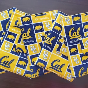May include: Four fabric items with a repeating pattern of yellow and navy blue squares. Each square features the text "Cal", "UC Berkeley", a bear, and the UC Berkeley logo. The items appear to be coasters or small pouches.