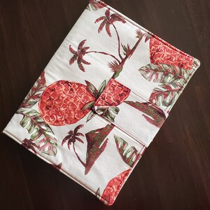 May include: A white fabric case with a tropical pattern featuring red pineapples, green leaves, and brown palm trees. The case has a flap closure and a stitched border. The overall design evokes a summery, vacation theme.