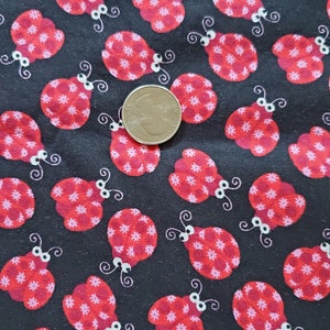 May include: Black fabric with a repeating pattern of red ladybugs with white spots and white eyes. The ladybugs are outlined in black. A US quarter is included for scale.