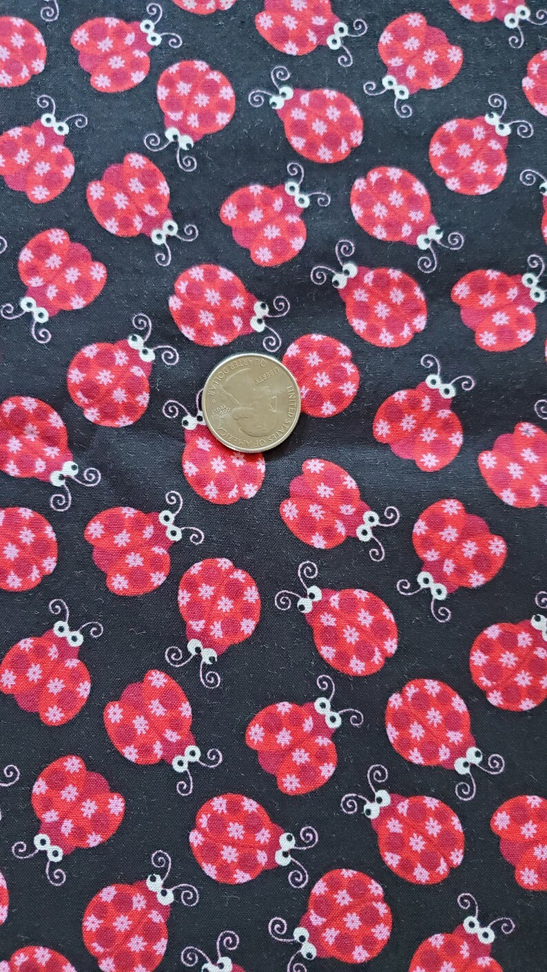 May include: Black fabric with a repeating pattern of red ladybugs with white spots and white eyes. The ladybugs are outlined in black. A US quarter is included for scale.