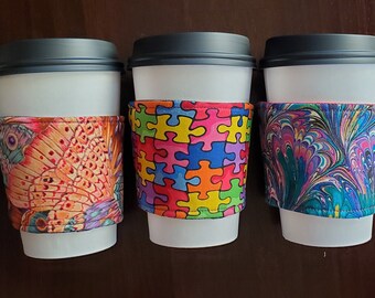 May include: Three white to-go coffee cups with black lids and colorful fabric sleeves. The sleeves feature a butterfly pattern, a puzzle piece pattern, and a marbled pattern.