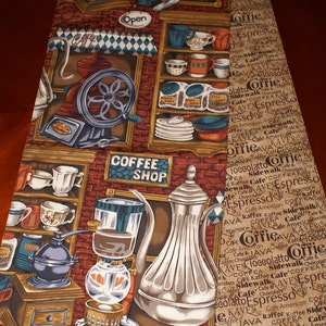 May include: A colorful fabric table runner with a coffee shop theme. The runner features a coffee grinder, a coffee pot, and a sign that says "Coffee Shop". The background is a brown brick pattern with a variety of coffee-related words printed on it.