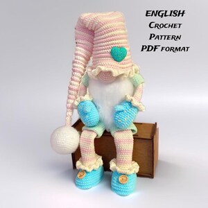 May include: A handmade crochet gnome doll with a long, striped pink and white hat, blue mittens and shoes, and a white beard. The gnome has a green heart on its hat and is sitting on a wooden box. Text on the image reads "ENGLISH CROCHET PATTERN PDF FORMAT."