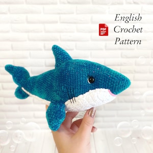 May include: A handmade, plush shark in shades of teal and white. The shark has a pink mouth, black eyes, and detailed stitching. The image includes the text "English Crochet Pattern".