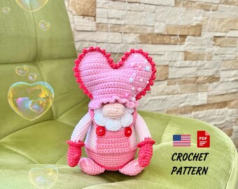 Könnte beinhalten: Eine handgefertigte gehäkelte Zwergenpuppe mit herzförmigem Hut und Latzhose. Die Puppe ist hauptsächlich rosa mit roten Akzenten und weißen Details. Das Bild enthält den Text "CROCHET PATTERN" und eine amerikanische Flagge.