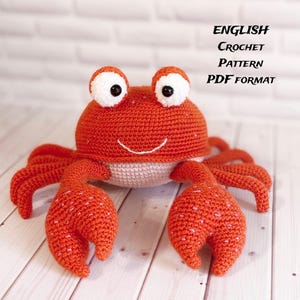 Pode incluir: Um caranguejo de crochê feito à mão em tons de laranja e branco. O caranguejo tem olhos grandes e expressivos, uma boca sorridente e garras detalhadas. O texto "ENGLISH CROCHET PATTERN PDF FORMAT" é exibido na imagem.