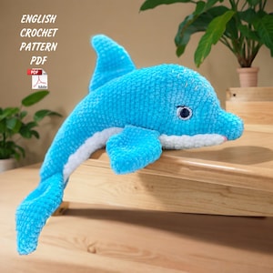 May include: A plush, turquoise crochet dolphin with white accents. The dolphin has a detailed eye and a pink mouth. The image also includes the text "ENGLISH CROCHET PATTERN PDF".