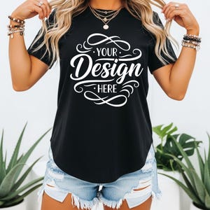 Puede incluir: Camiseta negra con el texto "YOUR Design HERE" en escritura blanca. La camiseta tiene un dobladillo redondeado y mangas cortas. La modelo lleva pantalones cortos vaqueros.
