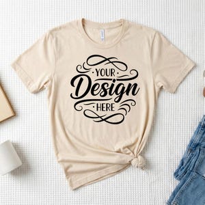 May include: A cream-coloured t-shirt featuring the text "YOUR Design HERE" in a black, decorative font. The t-shirt is knotted at the bottom and is displayed on a white, textured surface. The shirt has short sleeves and a crew neck.