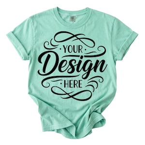 Puede incluir: Una camiseta verde menta con mangas enrolladas. La camiseta presenta el texto "YOUR Design HERE" en una fuente negra decorativa. El diseño está centrado en la parte delantera de la camiseta.