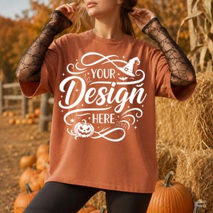Puede incluir: Una camiseta color óxido con un estampado gráfico blanco que dice "YOUR DESIGN HERE". El diseño incluye un sombrero de bruja, una calabaza, telarañas y murciélagos. Se lleva con mangas de encaje negro con un patrón de telaraña.