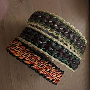K&ouml;nnte beinhalten: Zwei gewebte Armb&auml;nder. Eines hat ein Muster aus Orange, Rot und Schwarz. Das andere Armband hat ein Muster aus Braun, Gr&uuml;n und Beige. Beide Armb&auml;nder sind aus nat&uuml;rlichen Materialien gefertigt und f&uuml;r den t&auml;glichen Gebrauch geeignet.