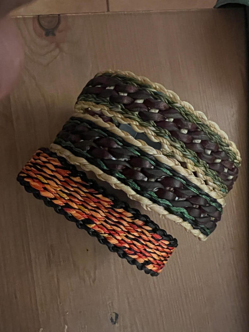 K&ouml;nnte beinhalten: Zwei gewebte Armb&auml;nder. Eines hat ein Muster aus Orange, Rot und Schwarz. Das andere Armband hat ein Muster aus Braun, Gr&uuml;n und Beige. Beide Armb&auml;nder sind aus nat&uuml;rlichen Materialien gefertigt und f&uuml;r den t&auml;glichen Gebrauch geeignet.