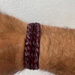 K&ouml;nnte beinhalten: Ein geflochtenes Lederarmband in Burgunder- und Lilat&ouml;nen. Das Armband hat ein gewebtes Design mit einer Kombination aus dunkelburgunderfarbenem Leder und helleren lilafarbenen F&auml;den. Das Armband ist etwa 2 cm breit.
