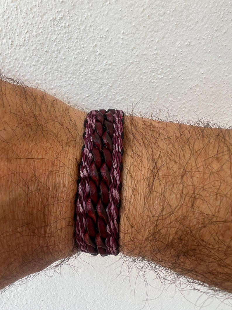 K&ouml;nnte beinhalten: Ein geflochtenes Lederarmband in Burgunder- und Lilat&ouml;nen. Das Armband hat ein gewebtes Design mit einer Kombination aus dunkelburgunderfarbenem Leder und helleren lilafarbenen F&auml;den. Das Armband ist etwa 2 cm breit.