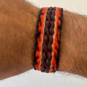 K&ouml;nnte beinhalten: Gewebtes Armband mit einem zentralen dunkelbraunen Band, das von leuchtend orangefarbenen und schwarzen R&auml;ndern flankiert wird. Das Armband wird an einem gebr&auml;unten Arm getragen und zeigt die kontrastierenden Farben und Texturen des Accessoires.