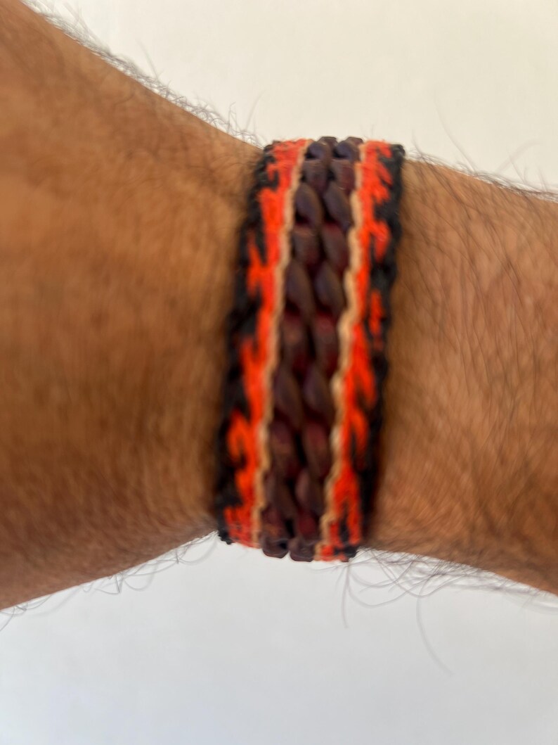 K&ouml;nnte beinhalten: Gewebtes Armband mit einem zentralen dunkelbraunen Band, das von leuchtend orangefarbenen und schwarzen R&auml;ndern flankiert wird. Das Armband wird an einem gebr&auml;unten Arm getragen und zeigt die kontrastierenden Farben und Texturen des Accessoires.
