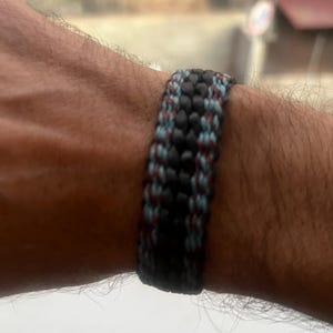 K&ouml;nnte beinhalten: Ein gewebtes Armband in Schwarz, Blau und Burgunderrot. Das Armband besteht aus einer Reihe von ineinander verwobenen Kordeln, wodurch eine strukturierte Oberfl&auml;che entsteht. Die Farben sind gleichm&auml;&szlig;ig verteilt.