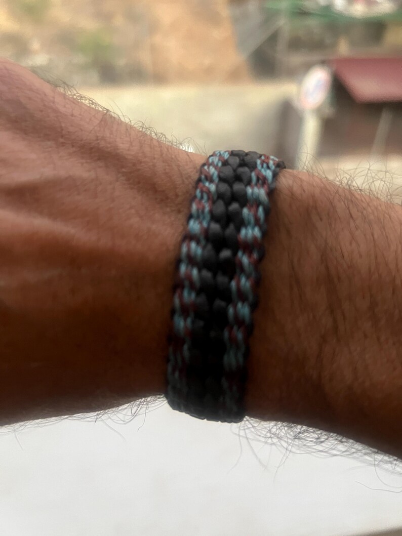 K&ouml;nnte beinhalten: Ein gewebtes Armband in Schwarz, Blau und Burgunderrot. Das Armband besteht aus einer Reihe von ineinander verwobenen Kordeln, wodurch eine strukturierte Oberfl&auml;che entsteht. Die Farben sind gleichm&auml;&szlig;ig verteilt.