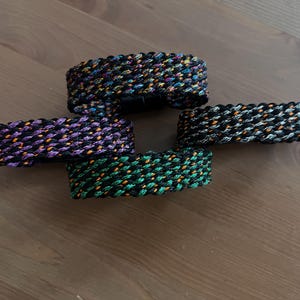 K&ouml;nnte beinhalten: Vier geflochtene Armb&auml;nder in verschiedenen Farben: Schwarz mit Orange und Gelb, Lila mit Orange, Schwarz mit Grau und Orange und Gr&uuml;n mit Orange. Die Armb&auml;nder sind aus einem gewebten Material.