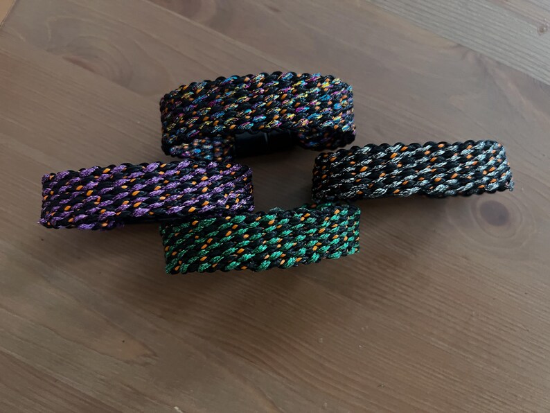 K&ouml;nnte beinhalten: Vier geflochtene Armb&auml;nder in verschiedenen Farben: Schwarz mit Orange und Gelb, Lila mit Orange, Schwarz mit Grau und Orange und Gr&uuml;n mit Orange. Die Armb&auml;nder sind aus einem gewebten Material.