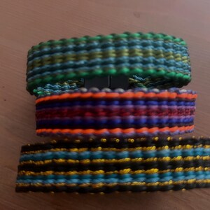 K&ouml;nnte beinhalten: Drei farbenfrohe gewebte Armb&auml;nder, die &uuml;bereinander gestapelt sind. Das obere Armband ist gr&uuml;n, blau und gelb. Das mittlere Armband ist orange, rot und lila. Das untere Armband ist schwarz, blau und gold.