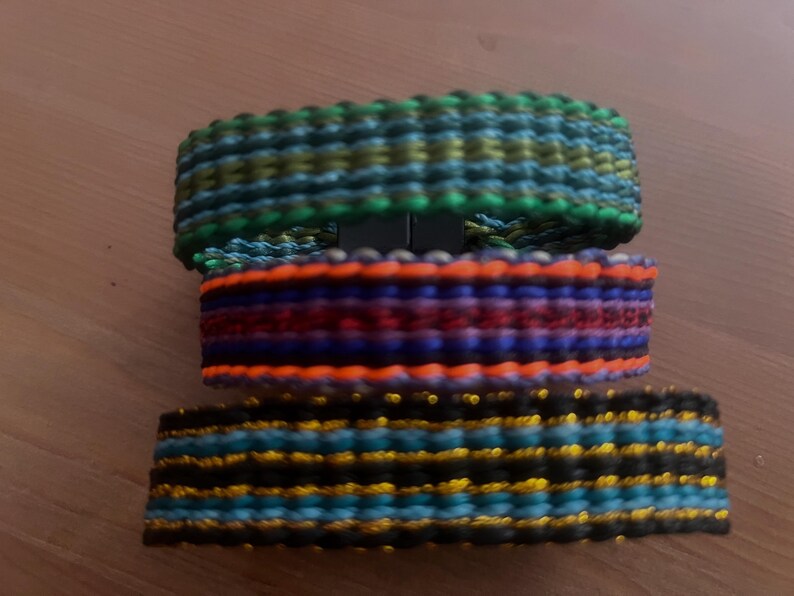 K&ouml;nnte beinhalten: Drei farbenfrohe gewebte Armb&auml;nder, die &uuml;bereinander gestapelt sind. Das obere Armband ist gr&uuml;n, blau und gelb. Das mittlere Armband ist orange, rot und lila. Das untere Armband ist schwarz, blau und gold.