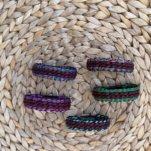 K&ouml;nnte beinhalten: F&uuml;nf geflochtene Armb&auml;nder in verschiedenen Farben, darunter Burgunderrot, Lila, Gr&uuml;n und Blau, werden auf einer gewebten Oberfl&auml;che pr&auml;sentiert. Jedes Armband hat einen Magnetverschluss. Die Armb&auml;nder sind etwa 2 cm breit.