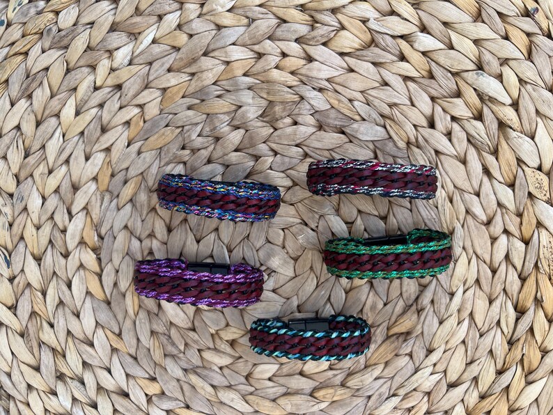 K&ouml;nnte beinhalten: F&uuml;nf geflochtene Armb&auml;nder in verschiedenen Farben, darunter Burgunderrot, Lila, Gr&uuml;n und Blau, werden auf einer gewebten Oberfl&auml;che pr&auml;sentiert. Jedes Armband hat einen Magnetverschluss. Die Armb&auml;nder sind etwa 2 cm breit.