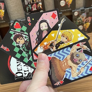 May include: A hand holding a collection of playing cards featuring characters from the anime series Demon Slayer. The cards have black backgrounds with character illustrations and suit symbols. The cards include the Ace of Hearts, Queen of Diamonds, and other face cards.