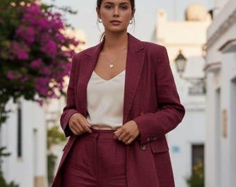 May include: A burgundy suit, consisting of a blazer and matching pants, paired with a cream-colored top. The blazer has a two-button closure. The outfit is worn outdoors, with a background of white buildings and purple flowers.