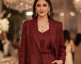 May include: A burgundy suit consisting of a blazer, camisole, and shorts. The double-breasted blazer has two rows of buttons. The camisole has a V-neck. The shorts have a pocket. The person is wearing gold earrings and a necklace with green pendants.
