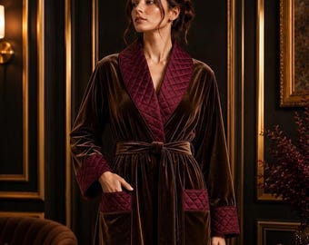 May include: A long-sleeved brown velvet robe featuring a quilted burgundy shawl collar, matching quilted burgundy cuffs, and two front pockets with quilted burgundy trim. The robe includes a matching velvet tie belt at the waist.