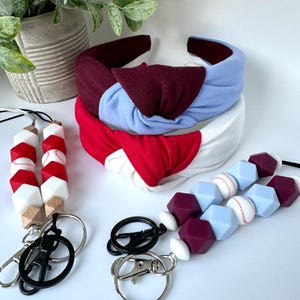 May include: Three headbands in red, white, and blue with a knotted design. Two silicone beaded keychains with baseball-themed charms. One keychain has white, red, and brown beads. The other keychain has white, light blue, and burgundy beads.