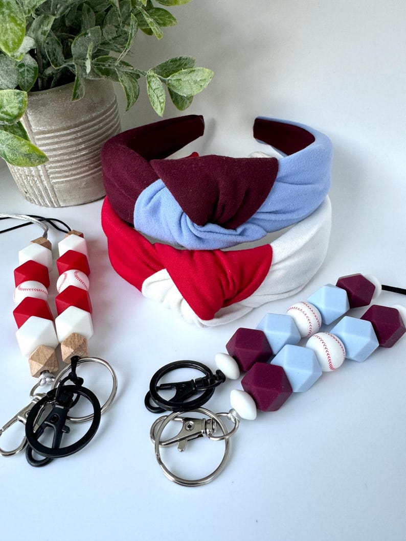 May include: Three headbands in red, white, and blue with a knotted design. Two silicone beaded keychains with baseball-themed charms. One keychain has white, red, and brown beads. The other keychain has white, light blue, and burgundy beads.