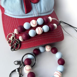 May include: A set of two beaded keychains with a baseball theme. The keychains are made with light blue and maroon beads, with white baseballs printed on some of the beads. The keychains are attached to silver key rings. A red and blue baseball cap with a red "P" on the front is in the background.